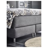 Modern grey upholstered bed with patterned decorative pillows, stylish stainless legs, and plush rug, exuding comfort and elegance.