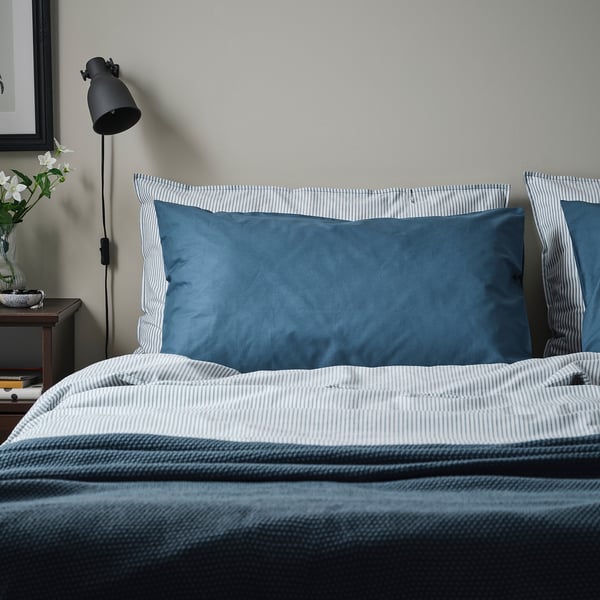 A bed with DVALA blue pillows. The pillows are rectangular, made of soft cotton, and fit neatly on the bed, contrasting with a striped bedding design.