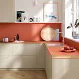 Modern kitchen with terracotta countertop, sink, wooden tray, and pomegranates. Beige cabinets, light floors, white walls.