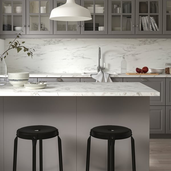 Modern kitchen with marble-patterned counter, grey cabinets, black stools, hanging lamp, and glass cabinet dishes.