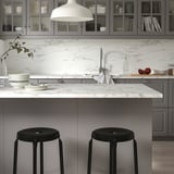 Modern kitchen with marble-patterned counter, grey cabinets, black stools, hanging lamp, and glass cabinet dishes.