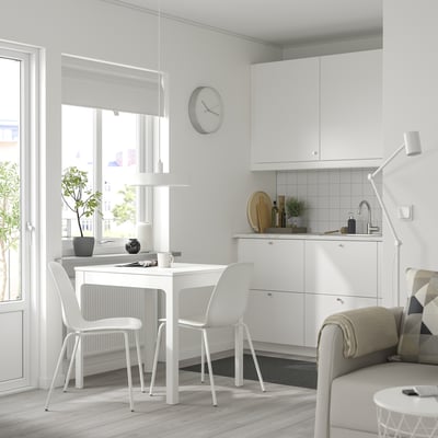 A small, modern living room with a white EKEDALEN extendable table, wooden chairs, a white kitchen with a sink, cupboards, and a window.