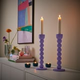Two tall purple candlesticks with lit candles on a white shelf. Small tealight candles are also present, creating a warm and inviting ambiance.