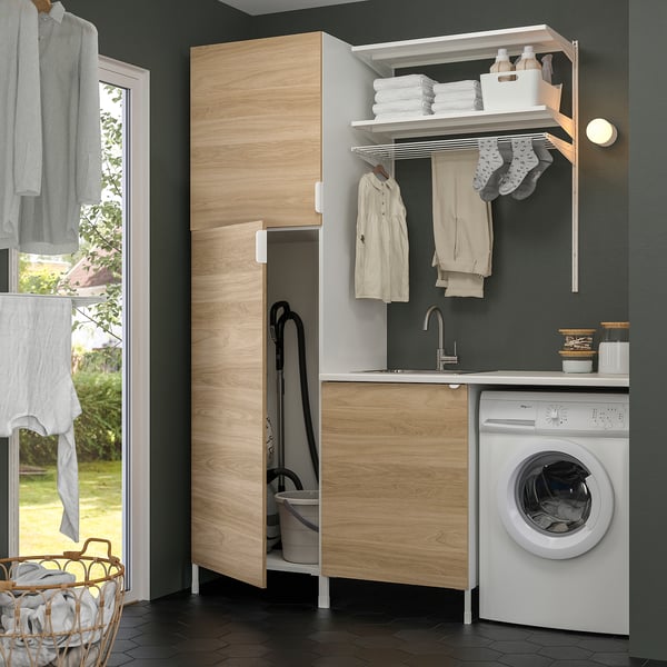 Modern laundry setup with ENHET wooden cabinet, sink, washing machine, and drying rack. Clothes hang and fold neatly.