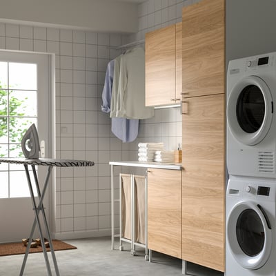 Modern laundry room with ENHET system. Wood-like cabinets, white appliances. Clothes and ironing board nearby.