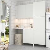 Modern white laundry room with ENHET cabinets, sink, washing machine, and dryer. Tiles, countertop, and cloth hamper visible.