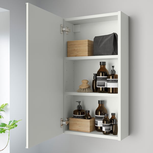 A modern bathroom cabinet with mirrored door open, revealing three shelves stocked with bottles, containers, and a hairbrush, mounted on a gray wall.