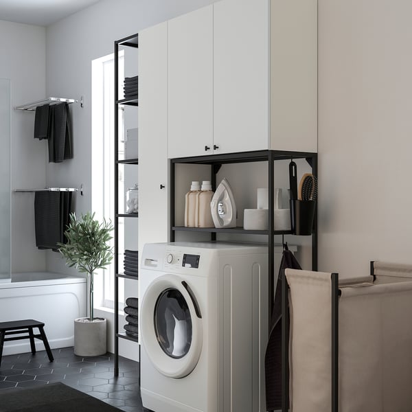 Modern laundry area with ENHET white units. Front-loading washer under black-framed shelves, towel rack to left, brown plant, black tools.
