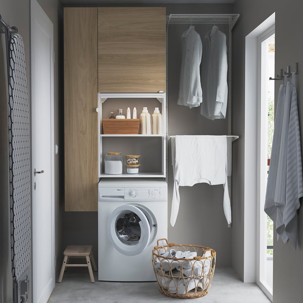 Modern laundry setup with ENHET series: white washing machine, wooden cabinet, hanging shirts, basket with towels.