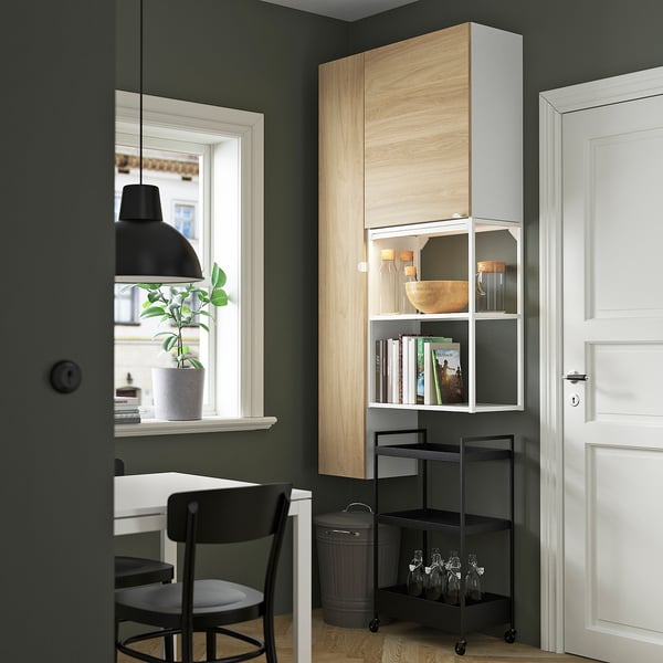 Modern room with ENHET storage. Tall unit, light wood, white shelves, black chair, plant, and bottles. Door open, space used efficiently.