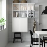 Modern kitchen with ENHET white cabinet & black stool, holding dishes & books, next to window with plant.