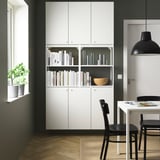 Modern white storage unit with shelves. Black chairs and pendant light. Green plant on window sill.