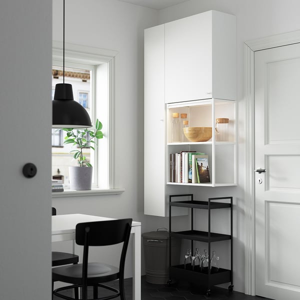 Modern room with white storage, black trolley, stool, & lamp. Natural light, plant.