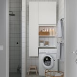 Modern bathroom with ENHET white storage cabinet, washing machine, hanging towels, wooden stool, and wicker laundry basket, next to shower.