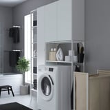 Modern laundry area with ENHET washer, storage, and rolling bag. Wall-mounted, black towels, plant.