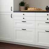 Modern white kitchen cabinets with gray countertop; includes potted plant and bread, drawers and cabinets with dark handles.