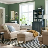 Cozy living room with green walls, beige sofa, striped rug, dark wooden cabinet, and large window.