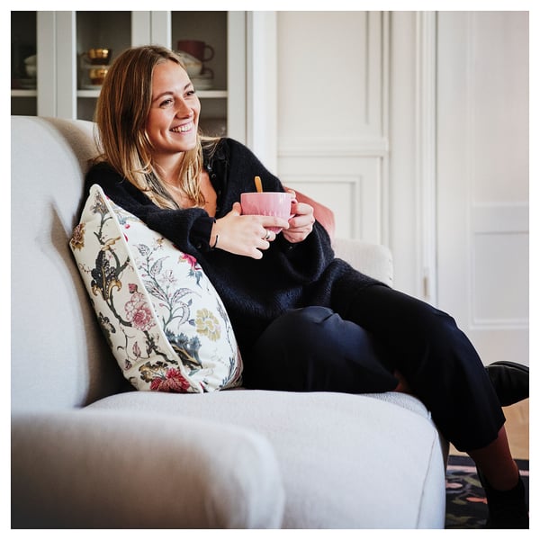 Woman relaxes on soft, white ESSEBODA sofa, holding pink mug, dressed in dark sweater and pants. Floral pillow behind her.