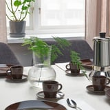 Table setting with FÄRGKLAR cups, saucers, and a vase with greenery; coffee maker, spoons, and plates nearby.