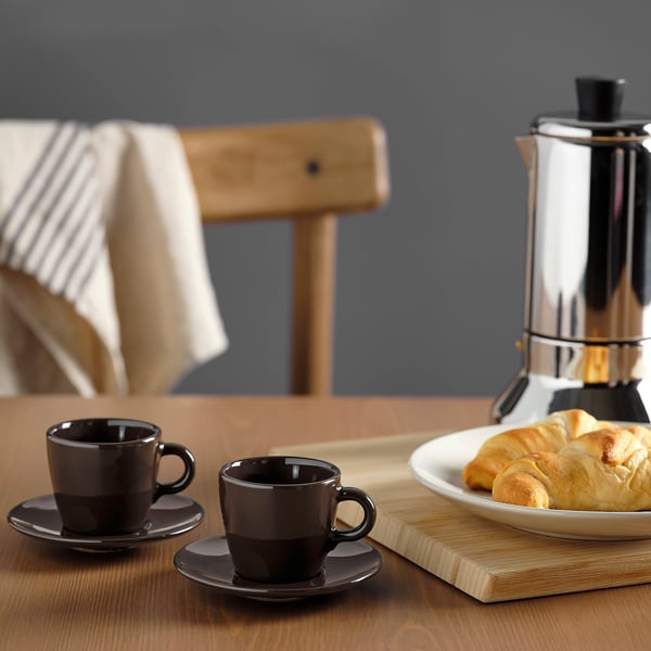 Two dark cups of coffee on saucers near a plate of pastries and a shiny coffee maker on a wooden table.