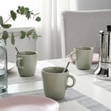 Two mugs with spoons on a table mat, alongside a water jug and coffee maker, indicating preparation for serving coffee or tea.