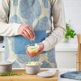 A person is grating cheese into a bowl using a glass bowl. They wear an apron and a wooden cutting board is underneath.