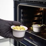 Hand wearing oven glove holding white oven-safe dish with baked food, ready to place inside black oven with another dish inside.