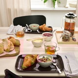 Table set with FÄRGKLAR plates, cups, and cutlery for breakfast. Includes croissants, granola, tea, and butter.