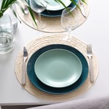 Table setting with teal plates and woven placemats. Wine glasses, cutlery, and a plant are included.