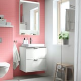 Small white bathroom mirror with light, mounted on pink-tiled wall, reflecting window and towels.