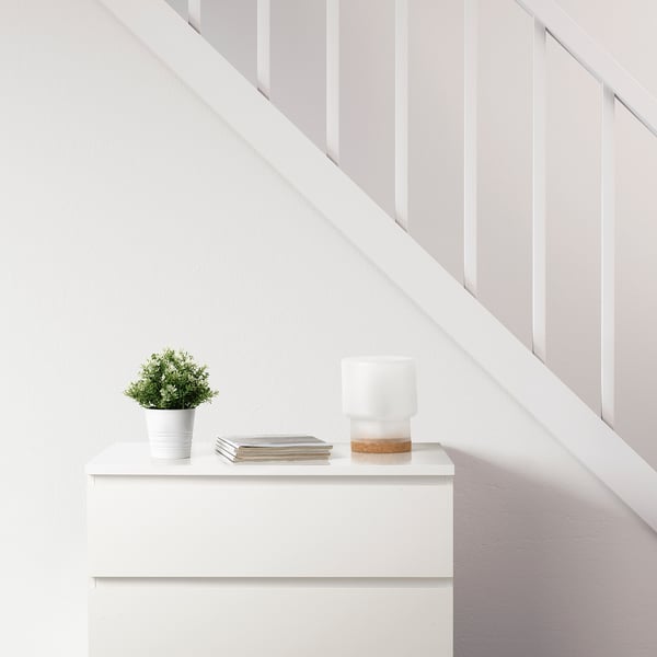 Minimalist room with white drawer, FEJKA plant, magazines, and lamp with cork base.