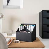 Office desk with organizer holding markers and pens, beside a roll of tape on a cork mat, next to a stack of drawers.
