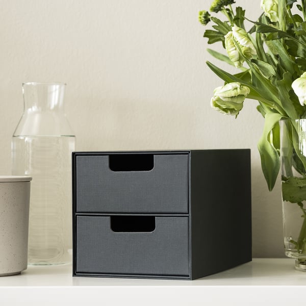 Black mini storage chest with two drawers on a white shelf, next to a vase of flowers and a glass jug.