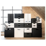 Stacked black and white storage boxes. Boxes vary in size, arranged neatly on wooden flooring and plywood wall. Modern and minimalist design.