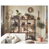 Modern living room with white couch, metal and wood shelving unit, potted plants, decorative vases, books, and wall hangings on beige walls.