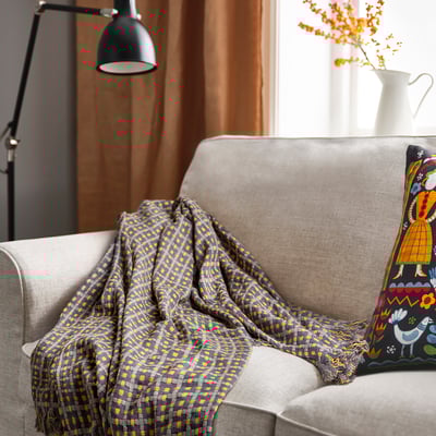 Grey/yellow FLIKBRUNÖRT blanket on sofa, with pillow & flowers.