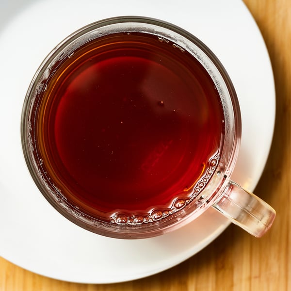 Clear glass mug filled with dark red tea on a white saucer. Calming, steaming beverage with rich flavor.