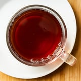 Clear glass mug filled with dark red tea on a white saucer. Calming, steaming beverage with rich flavor.