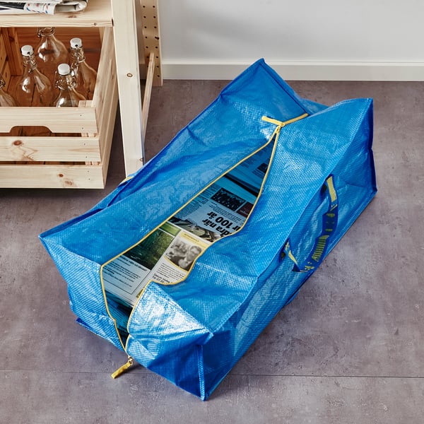 Blue storage bag with yellow handles and zipper, lying on grey floor next to wooden shelf.