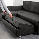 Person lifting seat of black FRIHETEN sofa bed revealing storage space.