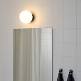 A round bathroom light fixture with a glass shade mounted above a mirror, providing general lighting. Two dark towels hang on chrome hooks beside it.