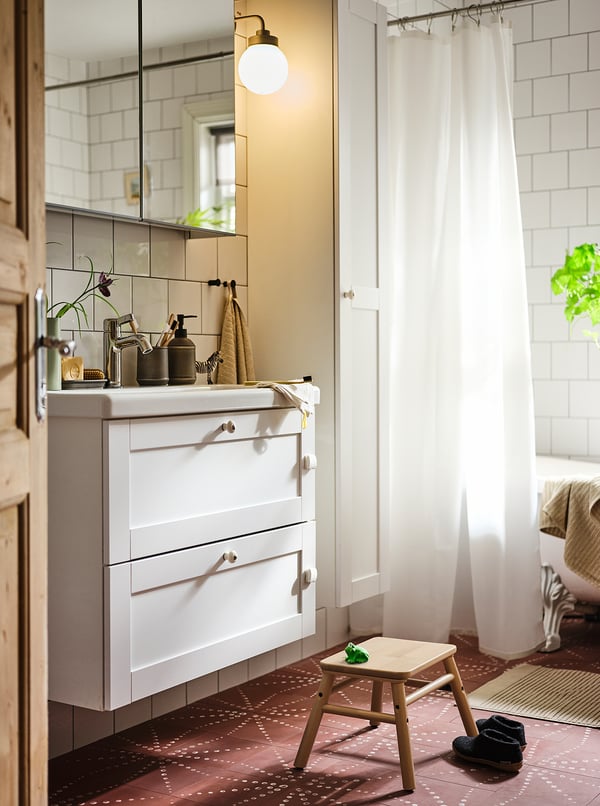 A well-lit bathroom with white tiles, a white vanity, and pinkish-brown patterned floor tiles. Wooden stool and a white shower curtain are visible.