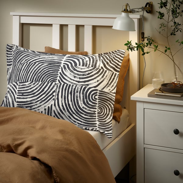 Bed with white FRÖDD pillowcase, printed black swirls, tan blanket, wooden headboard, wall lamp, nightstand with lamp, books, and glass.