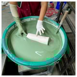 A person dips a white ceramic piece into a green glaze bucket using a wooden tool. The process creates a textured, bark-like design on the ceramic.