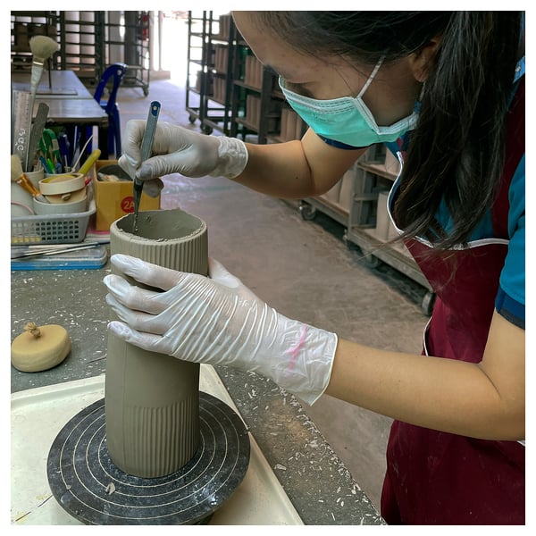 In a person is crafting a green, ribbed ceramic vase. Using a tool, they smooth the surface, with gloves and a mask on.