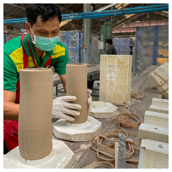 Man making earthy, wavy ceramic vases in cluttered studio. Alternate: man creates wavy ceramic vases, wearing colorful shirt and face mask.