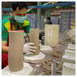 Man making earthy, wavy ceramic vases in cluttered studio. Alternate: man creates wavy ceramic vases, wearing colorful shirt and face mask.