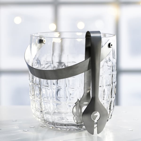 Clear textured glass ice bucket with attached stainless steel tongs and handle sits on a white surface.