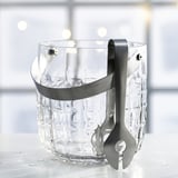 Clear textured glass ice bucket with attached stainless steel tongs and handle sits on a white surface.