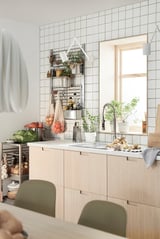Modern kitchen with light wooden cabinets, white tiles, green chairs, potted plants, and hanging vegetables.
